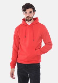 Felpa rossa con cappuccio, tasca frontale, cordini regolabili e polsini a costine. Abbinata a jeans scuri, presentata su uno sfondo bianco.