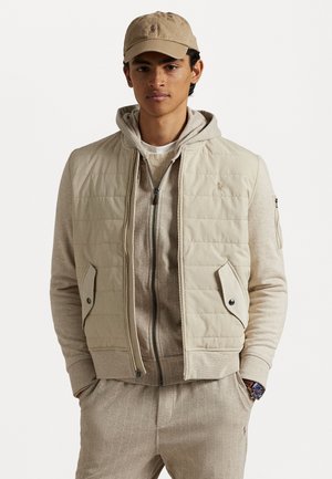 Beige quiltet vest med sidelommer over en lys grå hættetrøje. Modellen bærer en matchende beige cap og lys grå stribede sweatpants.