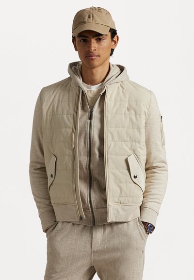 Beige quiltet vest med sidelommer over en lys grå hettegenser. Modellen har på seg en matchende beige caps og lys grå stripete sweatpants.