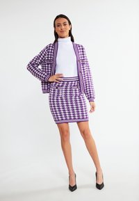 Violet houndstooth-kuvioinen neule, jossa on kultaiset napit, yhdistettynä samankaltaiseen hameeseen, molemmat valmistettu teksturoidusta kankaasta. Mustat korkokengät.