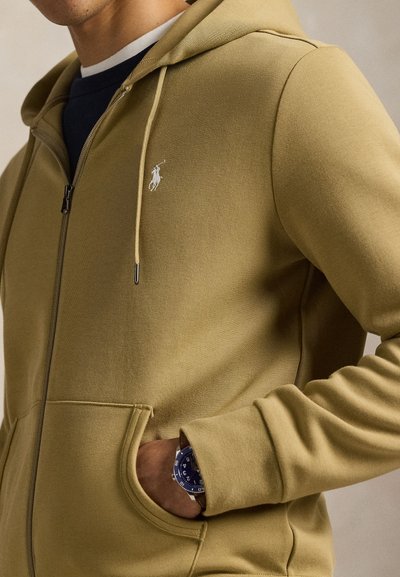 Χακί φούτερ με φερμουάρ, κατασκευασμένο από απαλό ύφασμα, με μικρό λευκό λογότυπο, τσέπες μπροστά και κουκούλα με κορδόνι. Συνοδεύεται από ένα ρολόι.