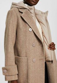 Beżowy płaszcz w jodełkę z odpinanym pikowanym kapturem, zapinany na pięć guzików, z bocznymi kieszeniami, o miękkiej fakturze i dopasowanym kroju.