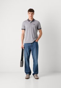 Γκρι και λευκό πουκάμισο πόλο με σχέδιο και σκούρο γκρι γιακά, συνδυασμένο με μπλε τζιν. Το μοντέλο κρατά μια γκρι τσάντα χιαστί με μαύρη λουρίδα.