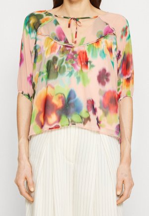 Frau trägt eine durchsichtige, mehrfarbige Blumenbluse mit einem Bindeband am Ausschnitt und Puffärmeln bis zum Ellbogen, kombiniert mit weißen, fein gestreiften Hosen.