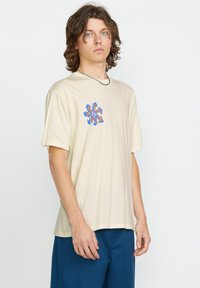 Camiseta de algodón beige con un estampado gráfico en azul y naranja en el lado izquierdo del pecho. Corte holgado, mangas cortas y cuello redondo.