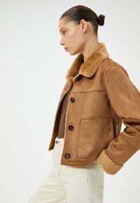 Veste en suède marron avec un col en fausse fourrure, dotée de trois grands boutons, de poches latérales et d'une texture douce. Portée avec un pantalon clair.
