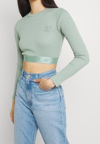 Kvinna i en mintgrön långärmad cropped topp med logo och elastisk kant, i kombination med ljusblå högmidjade jeans, med handen i fickan.