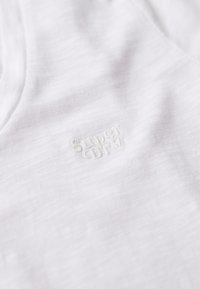 Tela de algodón blanco con una textura sutil, que presenta un pequeño logo bordado en color coincidente que dice "Super Dry".