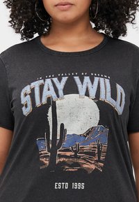 Černé grafické tričko s potiskem pouštní scenérie se kaktusy, horami a velkým měsícem. Text obsahuje "STAY WILD" a "ESTD 1995." Jemná textura.