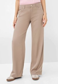 Beige weit geschnittene Hose mit glatter Textur, vorderen Taschen und einem Knopfverschluss. Kombiniert mit hellen Schuhen, die einen dezenten rosa Akzent aufweisen.