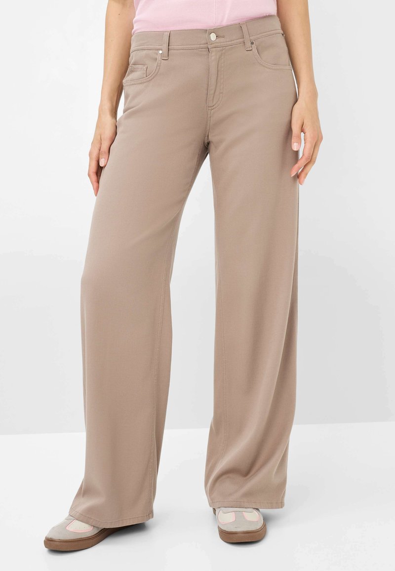 Beige weit geschnittene Hose mit glatter Textur, vorderen Taschen und einem Knopfverschluss. Kombiniert mit hellen Schuhen, die einen dezenten rosa Akzent aufweisen.