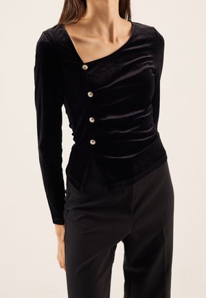 Femme portant un haut en velours noir à manches longues avec des boutons argentés en diagonale et un décolleté asymétrique, associé à un pantalon noir.