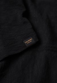 Zwart stoffen t-shirt met een gladde textuur en een klein oranje merklabel dat op de zijnaad is gestikt. Eenvoudig en minimalistisch ontwerp.