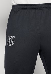 Sorte træningsbukser med elastisk talje, der har et hvidt broderet FC Barcelona-logo på venstre lår. Glat, tekstureret stof.