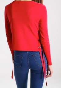 Červený top s dlouhými rukávy s cropped designem, ozdobený kovovými kroužky a vázacími detaily po stranách, kombinovaný s modrými džíny. Hladká textura látky.
