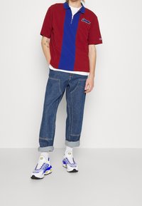 Personne portant un polo à manches courtes rouge et bleu, un jean bleu retroussé, des chaussettes blanches et des baskets blanches avec des accents bleus et jaunes, debout contre un mur blanc.