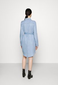 Kvinna med mörkt hår i en låg hästsvans, klädd i en bälte i ljusblå denimklänning och svarta ankelstövlar, som står mot en enfärgad bakgrund.