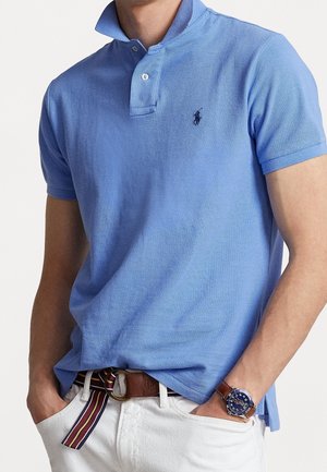 Lichtblauwe polo met een klassieke kraag, met een klein logo op de borst, gecombineerd met witte broek en een veelkleurige gestreepte riem.