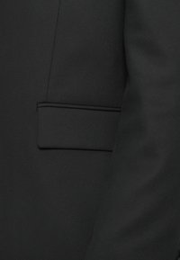 Secção de casaco preto com um bolso com piping subtil, textura de tecido suave e design estruturado.