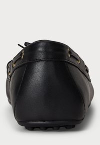 Μαύρο δερμάτινο μοκασίνι με θέα από την πίσω πλευρά, διαθέτοντας ραμμένα ραφάκια και χρυσές οπές με ορατή καουτσούκ σόλα στο κάτω μέρος.