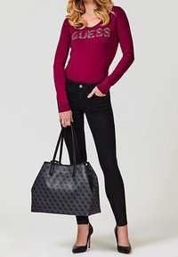Kobieta ubrana w bordową bluzkę Guess, czarne wąskie dżinsy, czarne szpilki, trzymająca dużą, wzorzystą czarną torbę na ramię.