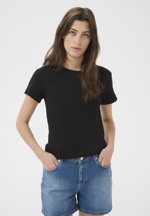 Jeune femme aux longs cheveux bruns portant un t-shirt noir uni à manches courtes et un short en denim bleu, les mains dans les poches, debout devant un fond blanc.