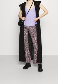 Lång svart stickad väst över en lavendelfärgad top med hålmönster, kombinerad med blommiga utsvängda byxor och svarta stövlar. Liten svart axelremsväska.