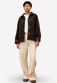 Femme debout portant une veste en peluche brun foncé, un haut blanc, un pantalon large beige et des chaussures à imprimé léopard devant un fond uni.