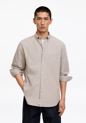 Jeune homme debout, une main dans la poche, portant une chemise beige boutonnée avec les manches retroussées et un pantalon foncé sur fond blanc.