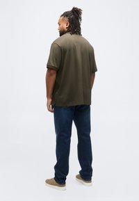 Άνδρας με dreadlocks στέκεται πλάτη, φορώντας πράσινο ελιάς πόλο πουκάμισο, σκούρα μπλε τζιν και μπεζ αθλητικά παπούτσια σε λευκό φόντο.