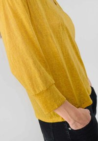 Haut jaune texturé avec une coupe décontractée et des manches trois-quarts. Présente un décolleté plongeant et une poche sur le côté, associé à un pantalon noir.
