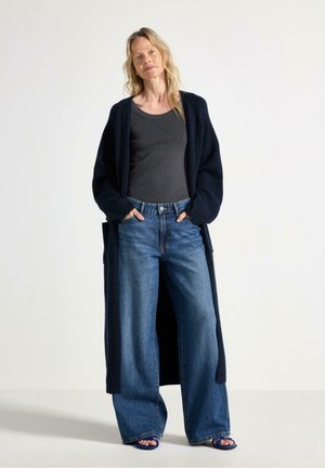 Donna con lunghi capelli biondi che indossa un top nero, un lungo cardigan scuro, jeans larghi blu e sandali blu con punta aperta, in piedi con le mani nelle tasche.