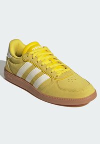 Gula sneakers med mockaliknande yta, vita tre ränder och en gummisul. Har en vadderad krage och perforerat tåområde.