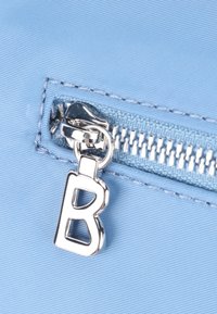 Ljusblått tyg med en silverblixtlås, med en metallisk bokstav "B" som dragkedjehandtag. Slät textur och rena sömnadsdetaljer.
