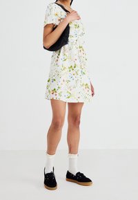 Robe fleurie à manches courtes, fond blanc, motifs verts et colorés. Sac tissé noir, chaussettes blanches à volants et chaussures noires.