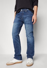 Hombre vistiendo pantalones vaqueros azules, zapatillas blancas con detalles beige y un suéter negro de manga larga, contra un fondo claro sencillo.