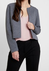 Chaqueta gris corta con textura de canalé, cuello redondo y sin cierre, llevada sobre una blusa rosa claro y pantalones negros.