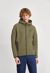 Olive green hoodie with a zip front, side zip pockets, and a black logo on the chest. Features a hood and textured fabric.