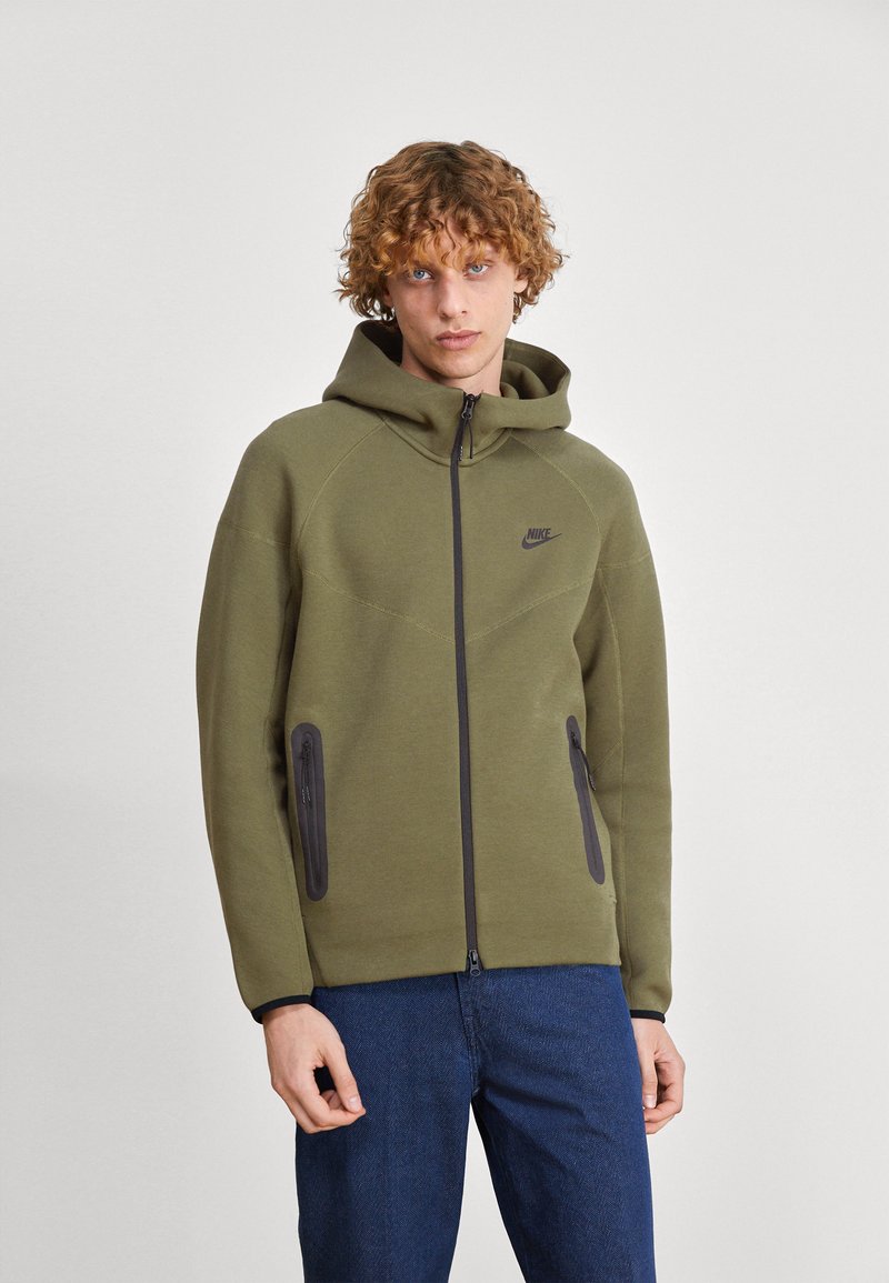 Olive green hoodie with a zip front, side zip pockets, and a black logo on the chest. Features a hood and textured fabric.