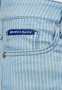 Lichtblauwe denim met witte verticale strepen, met een zaklabel in donkerblauw. Inclusief metalen knop en stikseldetails.