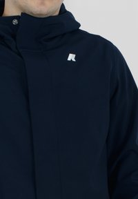 Tmavomodrá softshell bunda s vysokým golierom a malým logom na hrudi. Vyrobená z hladkého materiálu s elegantným dizajnom.
