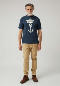 Hombre de mediana edad con cabello y barba canosos, vistiendo una camiseta de color azul marino con un diseño de ancla blanca, pantalones beige y botas marrones, de pie contra un fondo liso.