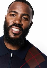 Námornícky zipsový sveter s veľkým kostkovaným vzorom, s hnedými a červenými akcentmi. Model má bradu a ľahký úsmev, na bielom pozadí.