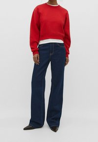 Femme portant un sweat-shirt rouge court, un sous-pull blanc, un pantalon large en jean bleu foncé et des chaussures noires pointues sur fond uni.