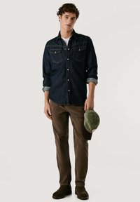 Camisa de mezclilla oscura con botones, dos bolsillos en el pecho, mangas arremangadas, combinada con pantalones de pana marrones y botas negras de tobillo. Gorro verde sostenido en la mano.