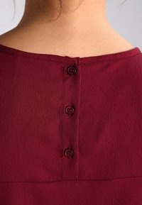 Bordeauxbluse med struktureret stof, bred halsudskæring og tre knapper på ryggen. Knapperne er runde og matcher stoffarven.
