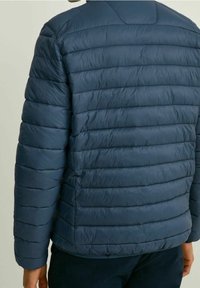 Veste matelassée bleu marine avec sections matelassées, texture lisse et col arrondi. Le design présente une silhouette épurée et des poignets élastiques.