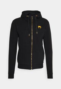 Černá mikina na zip vyrobená z měkké tkaniny, s zlatým zipem, žlutými akcenty na šňůrkách a malým zlatým logem na hrudi.