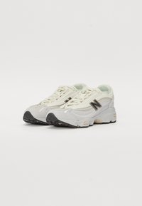 Zapatillas gris claro con parte superior de malla y ante, suela de goma, cordones blancos y detalles en negro, que presentan un destacado logotipo "N" en el lateral.