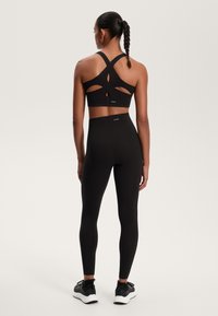 Črn športni top s krojenjem na hrbtni strani in izrezom, v kombinaciji z visokim pasom črnimi leginami in črnimi športnimi čevlji.
